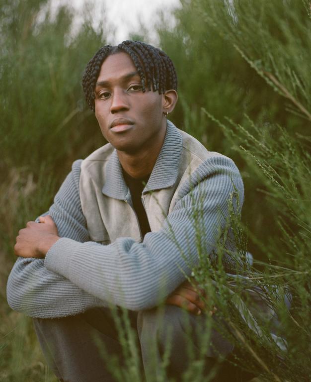 A man with braided hair crouches in a field of green plants.