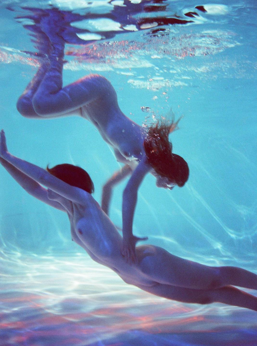 Two women swim nude underwater in a pool with light reflections on the surface.