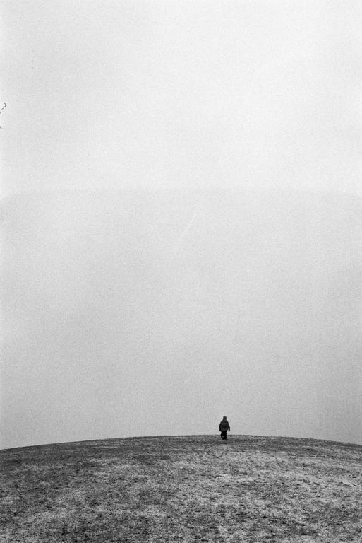 A lone figure walks away from the viewer over a hill under a vast, hazy sky.