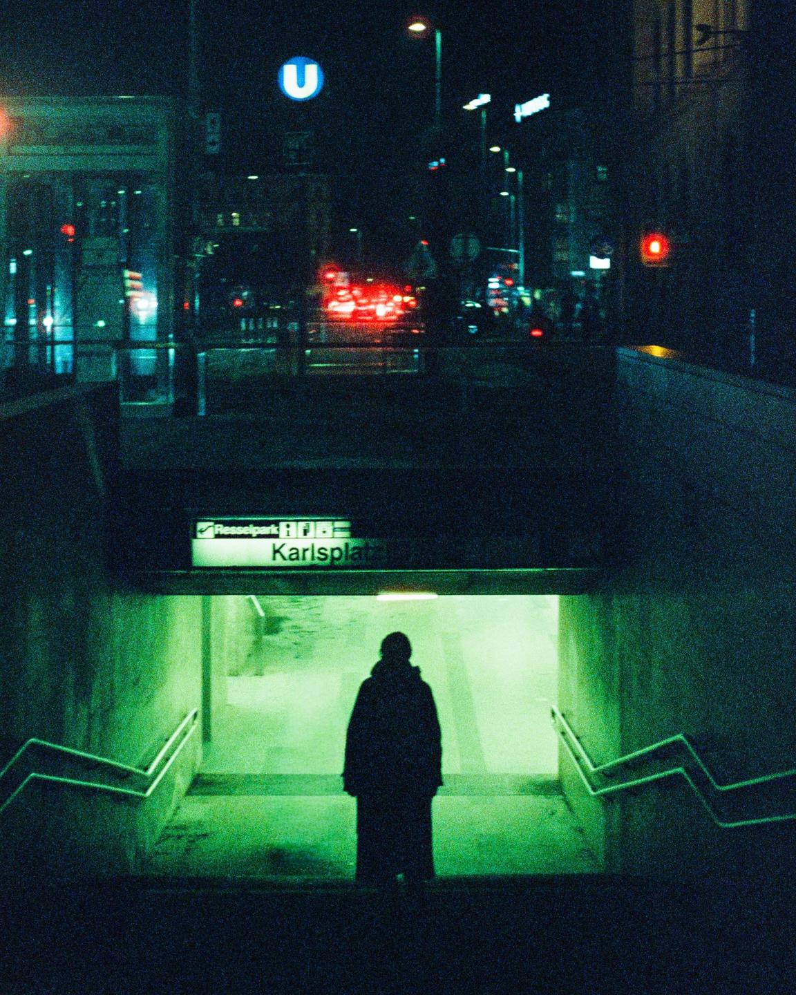A person stands at the top of a subway entrance with a street and traffic visible in the background at night.