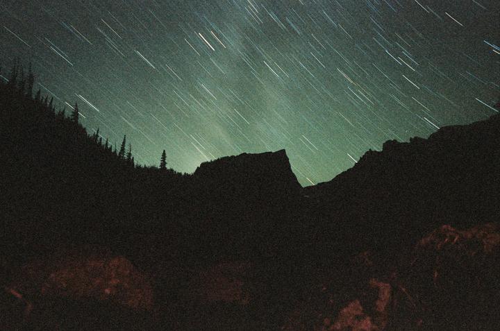 A long exposure photograph captures star trails above a dark mountain landscape with silhouetted trees.