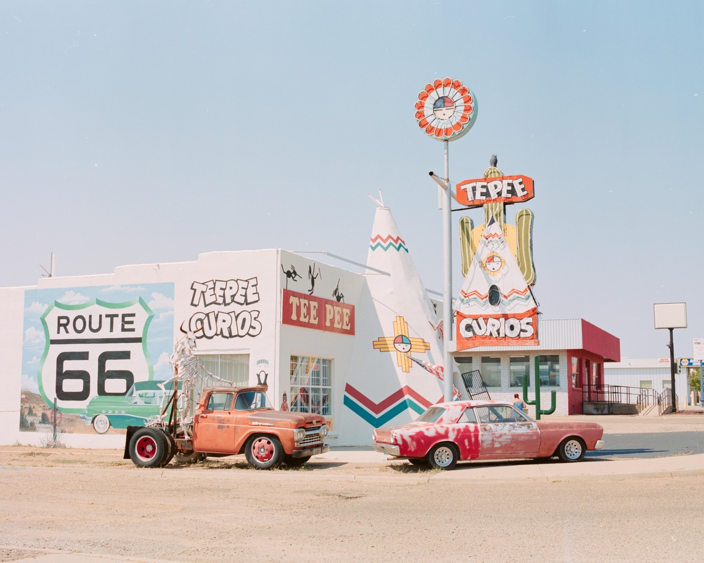 Two vintage vehicles are parked in front of a building with a Route 66 mural and a large teepee sign.