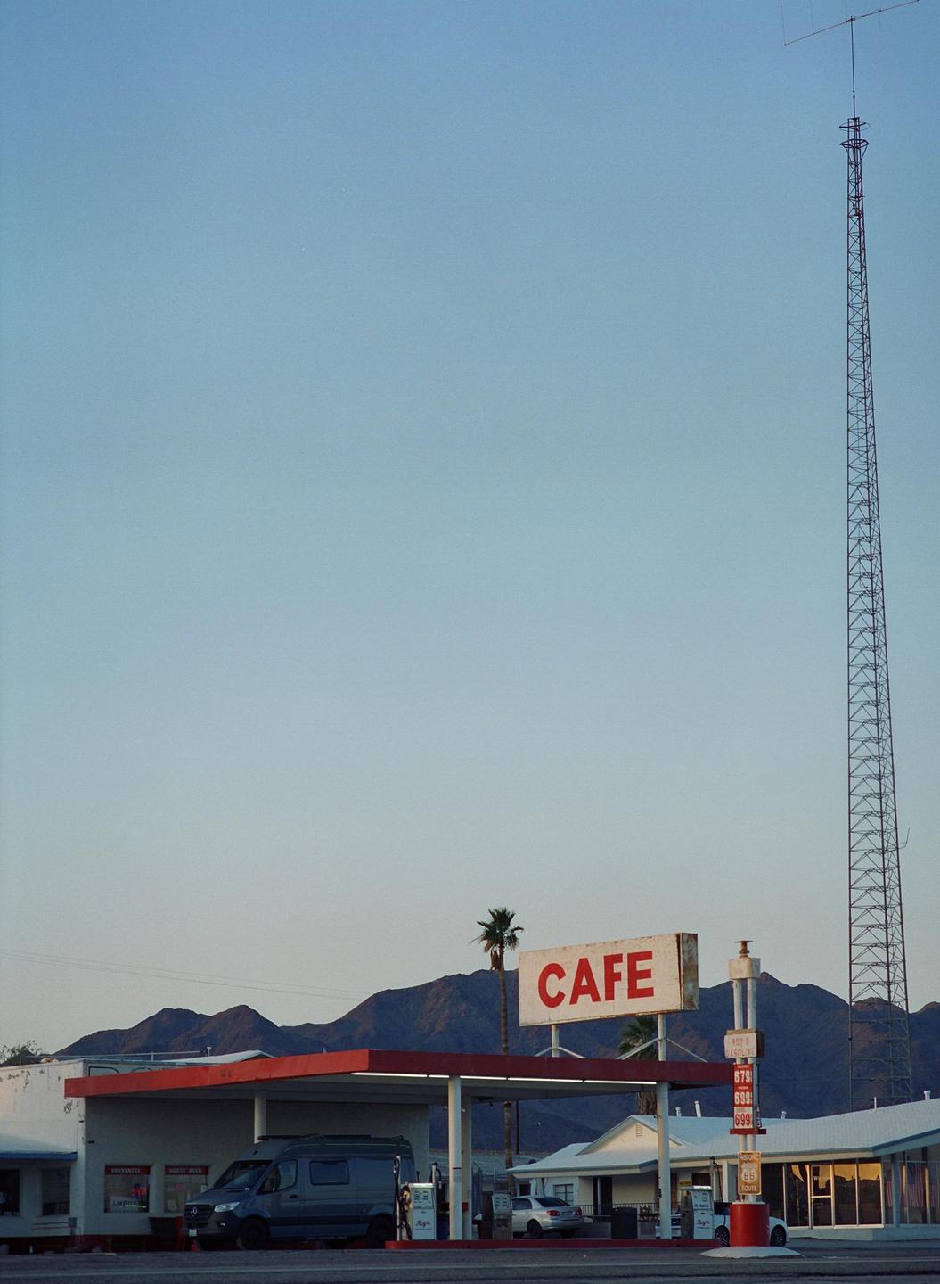A gas station with a "CAFE" sign stands in front of mountains and a tall antenna.