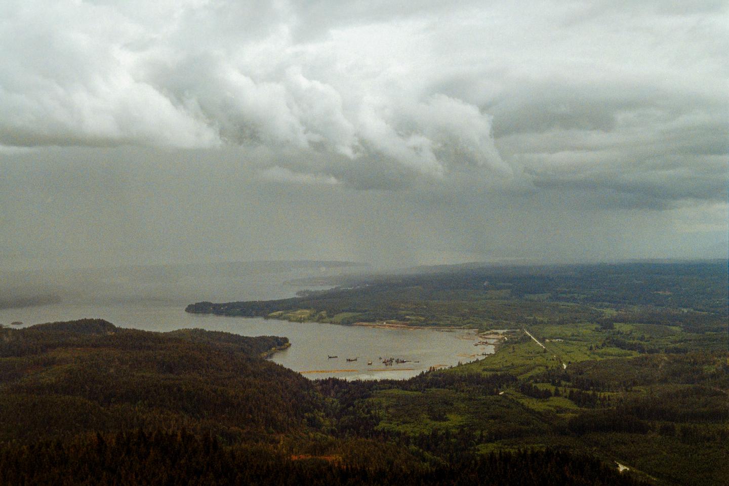 A wide landscape shows a body of water surrounded by forests under a cloudy sky with rain falling in the distance.