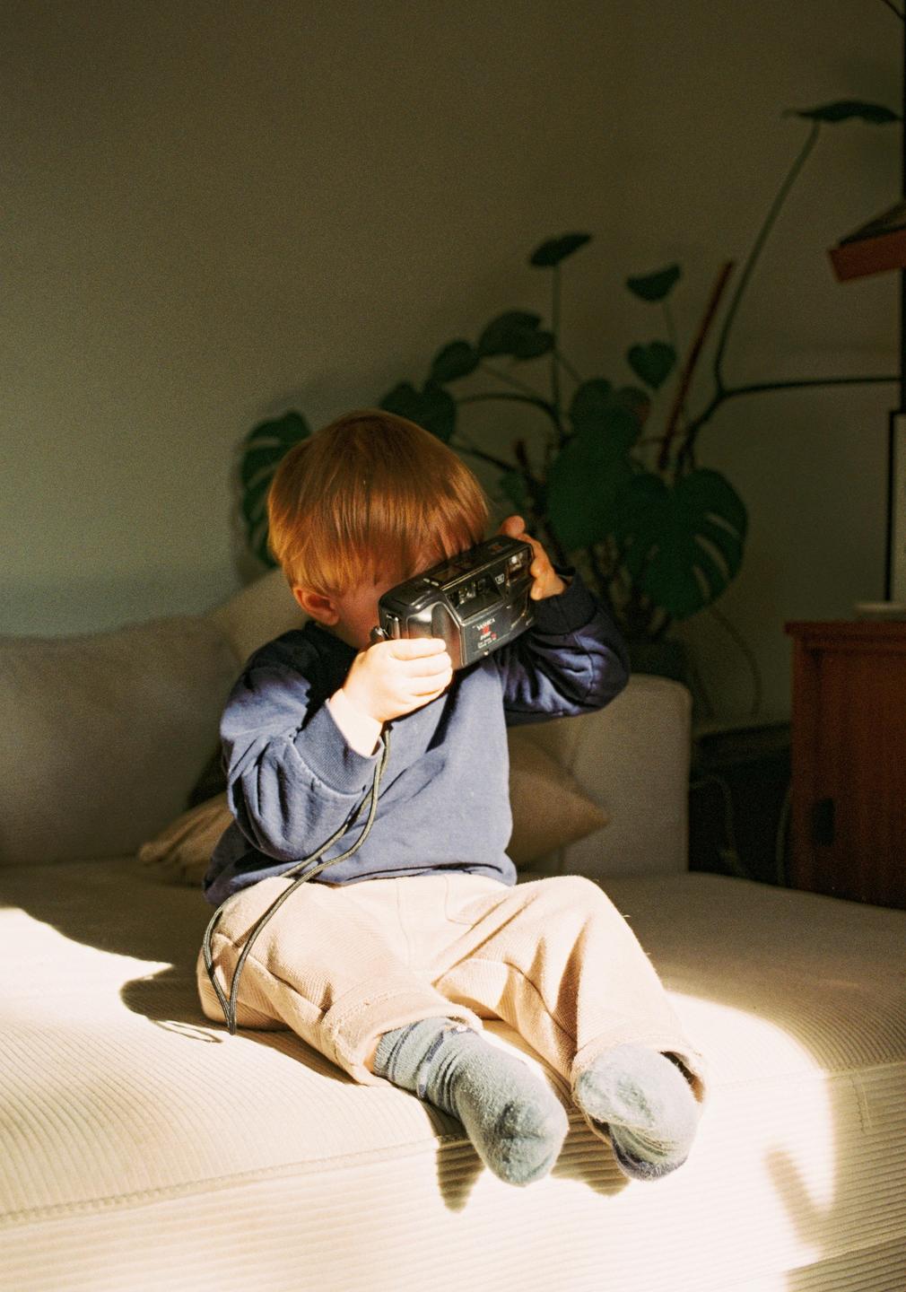 A young boy sits on a couch and holds a camera up to his face.