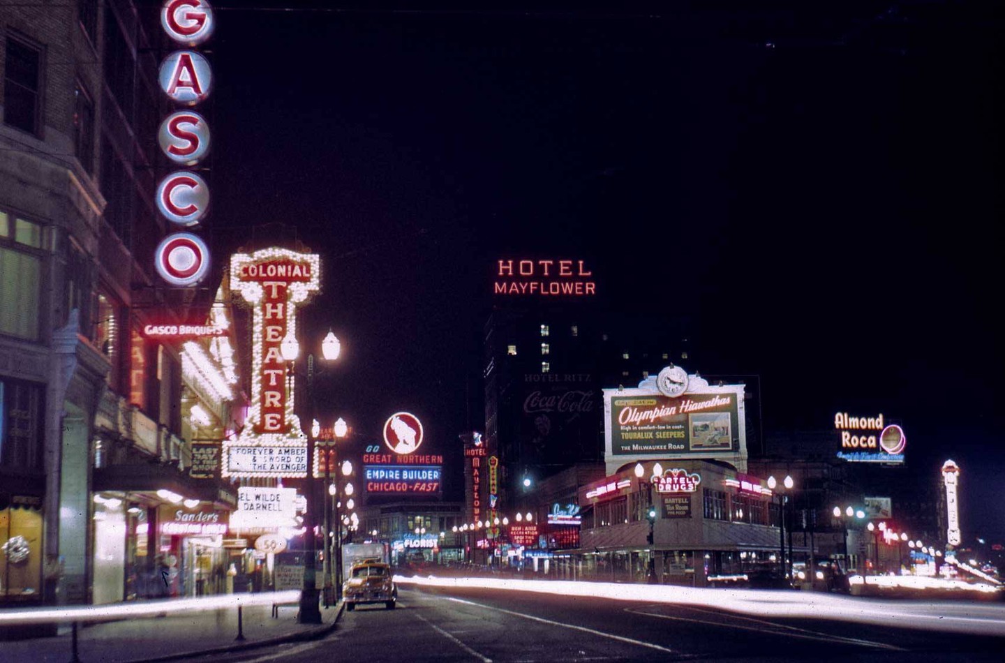 A street at night is illuminated by numerous neon signs and light trails from vehicles.