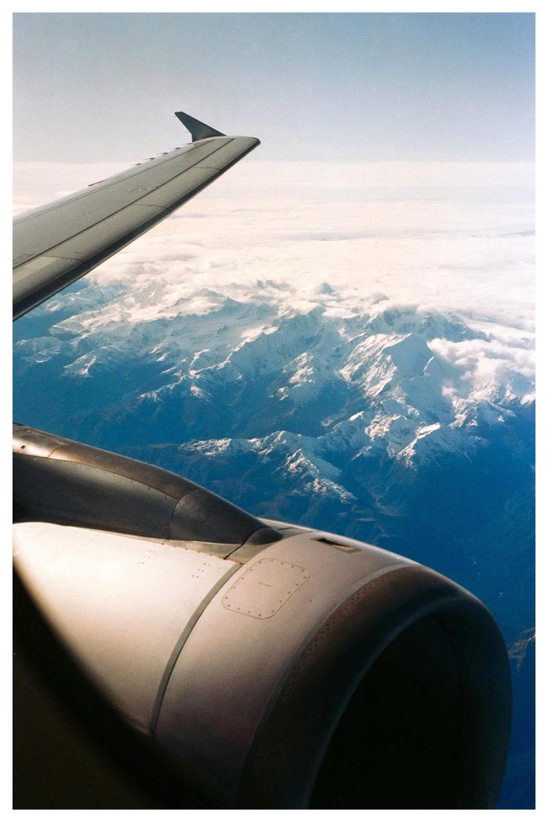An airplane wing and engine are visible above snow-capped mountains and clouds.