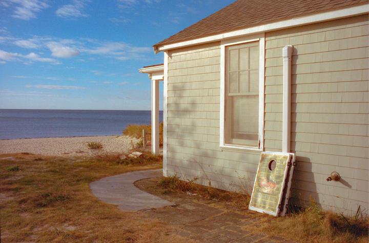 A house with a cornhole game board leaning against it sits next to a sandy beach and the ocean under a blue sky.