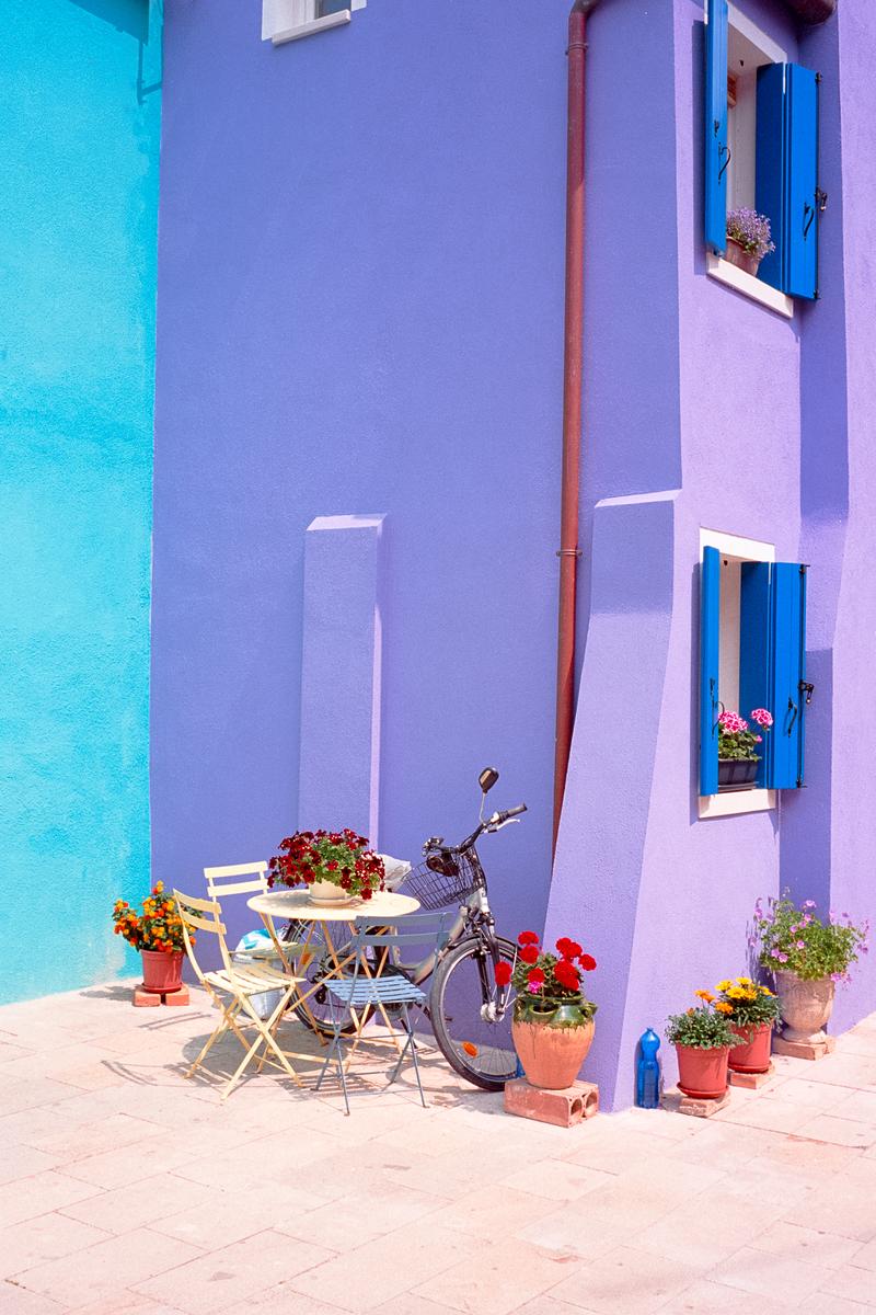 A bicycle is parked next to a table and chairs outside a purple building with blue shutters.