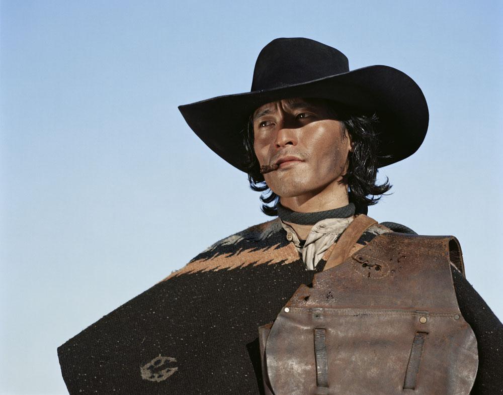 A man wearing a cowboy hat and poncho smokes a cigar and looks to the left.