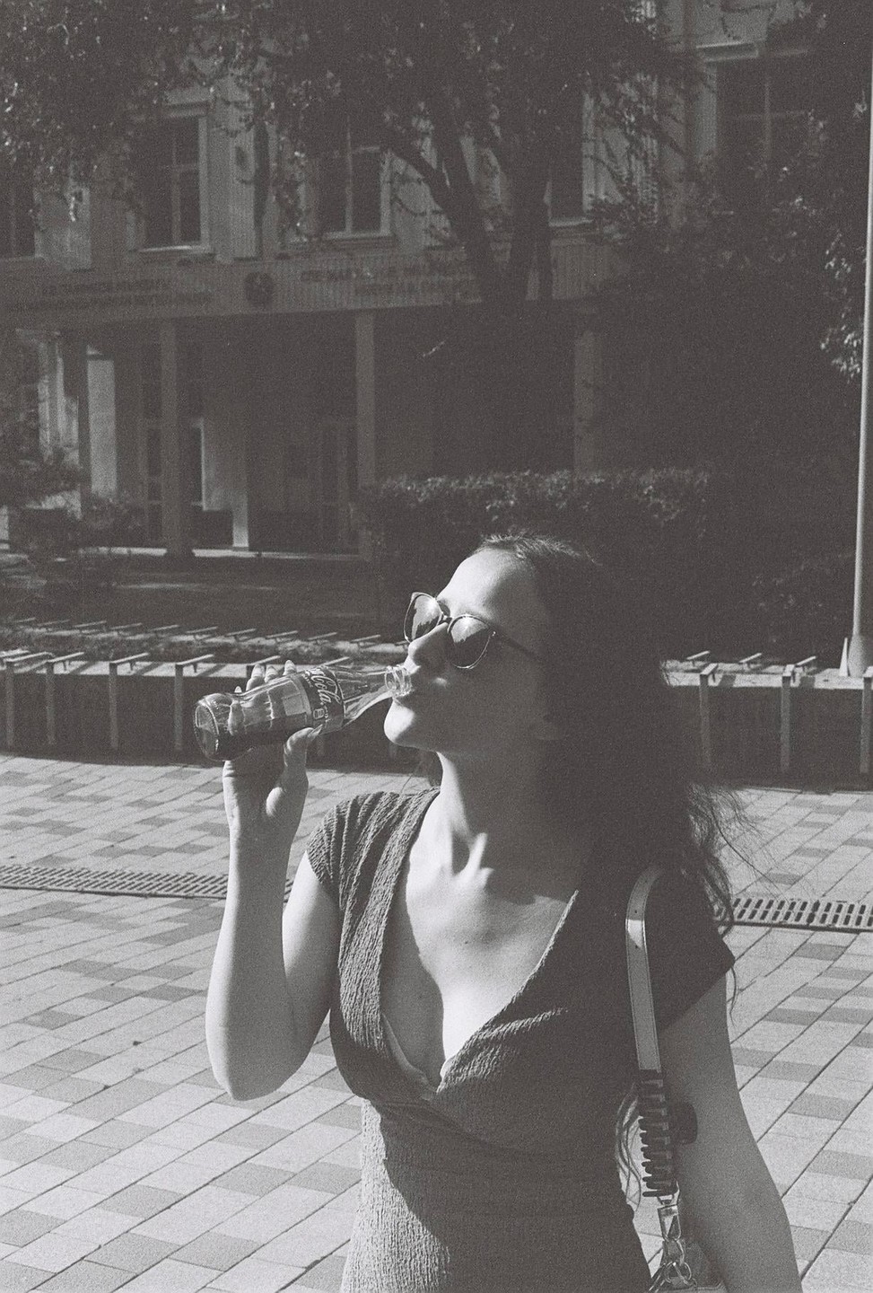 A woman wearing sunglasses drinks from a glass bottle on a paved surface in front of a building.