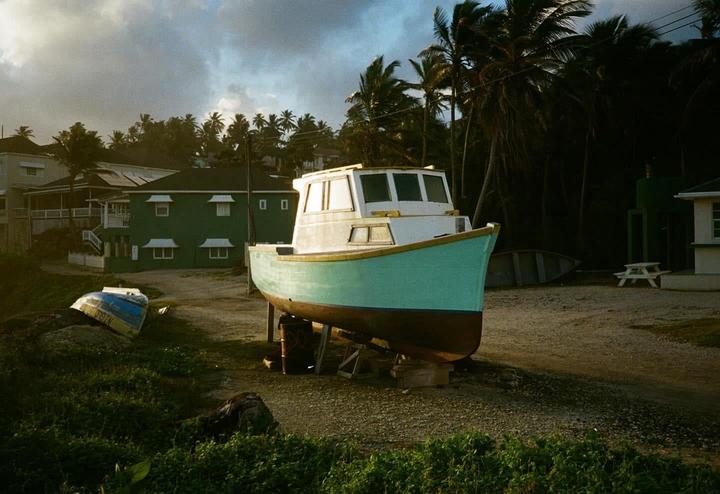 A turquoise and white boat sits on dry land with palm trees and buildings in the background under a cloudy sky.
