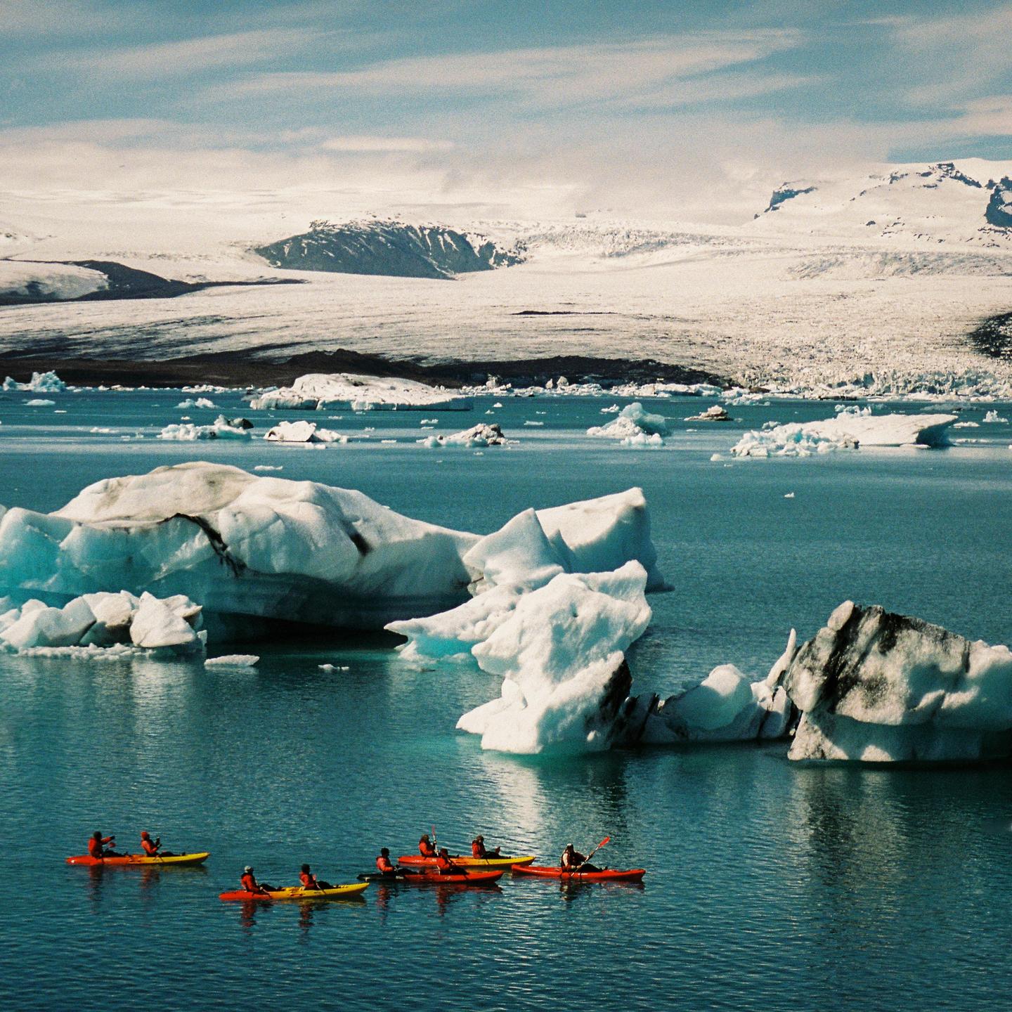 Several people kayak on a glacial lagoon with icebergs and snow-covered mountains in the background.