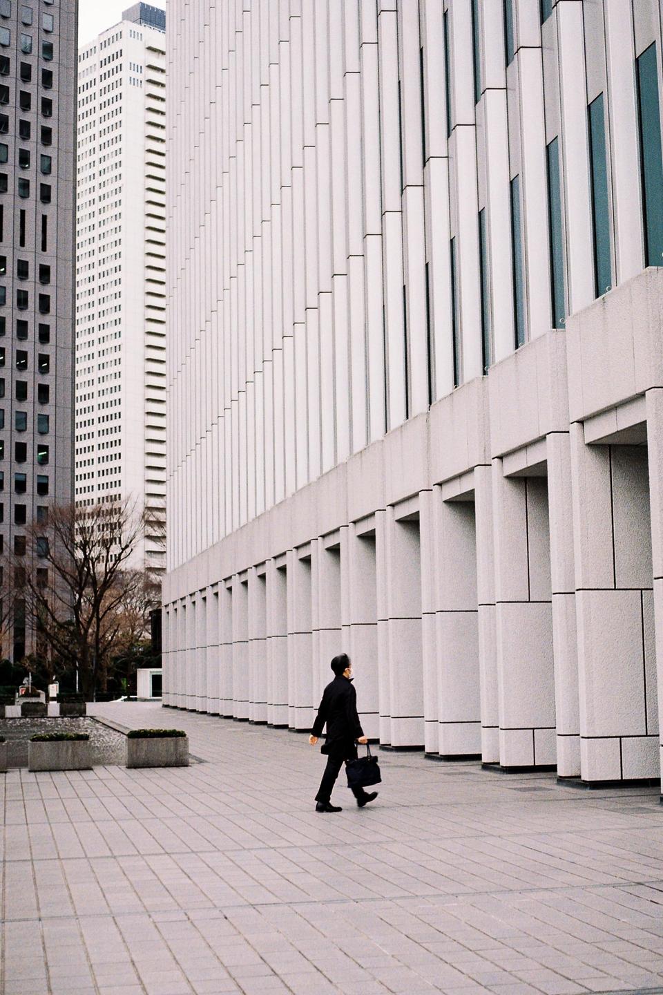 A man walks along a paved path next to a modern building with tall, narrow windows.