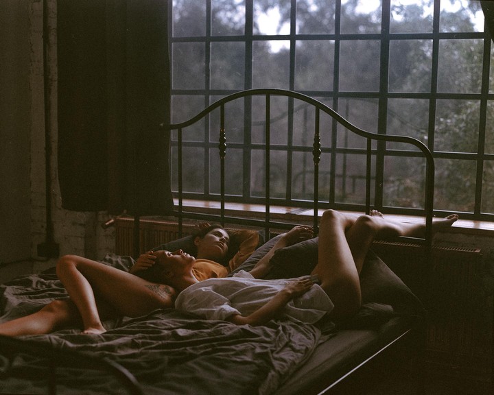 Two women are lying on a bed next to a large window with a view of trees.