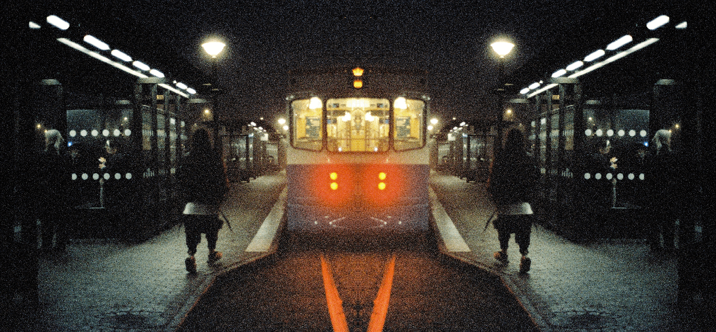 A person stands on a dark subway platform as a train approaches, its headlights glowing red.