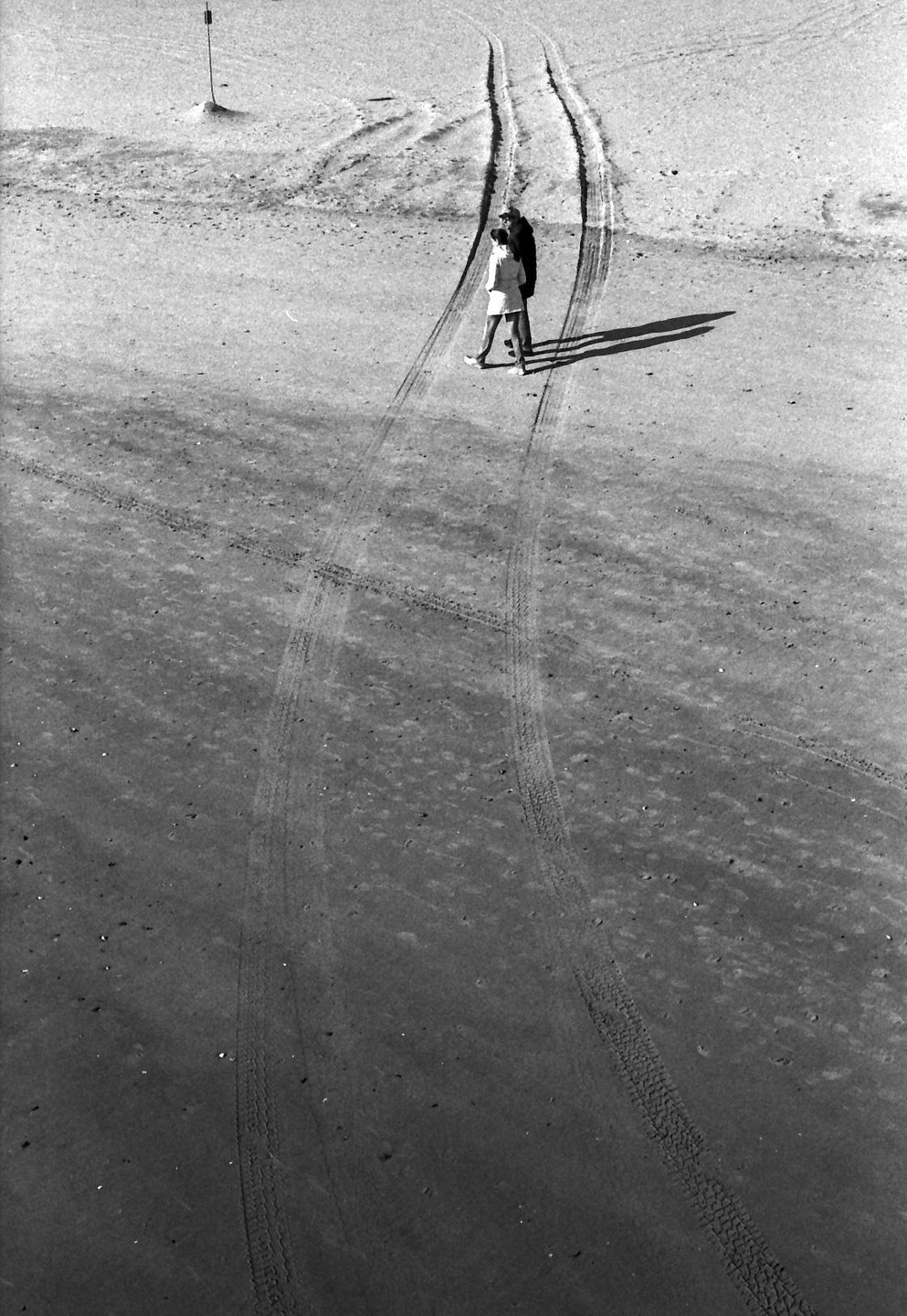Two people walk on a sandy surface with tire tracks and long shadows.