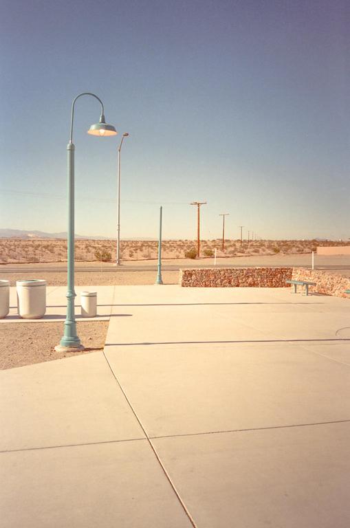 A light blue street lamp stands in the foreground of a desert landscape with a road, power lines, and mountains in the distance.