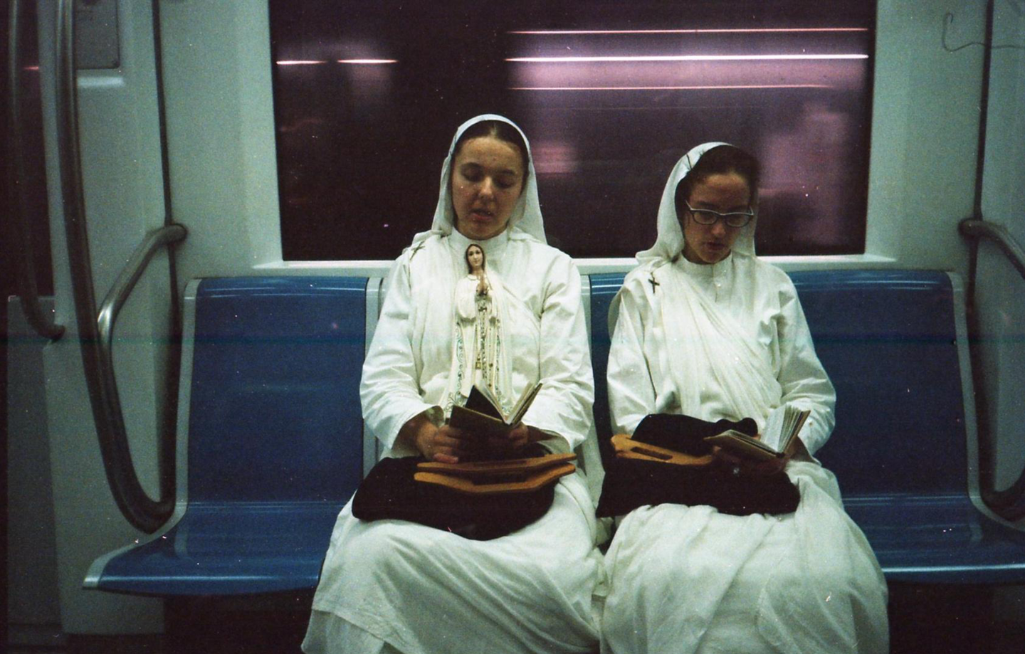 Two women in white habits sit on a subway train, reading books.
