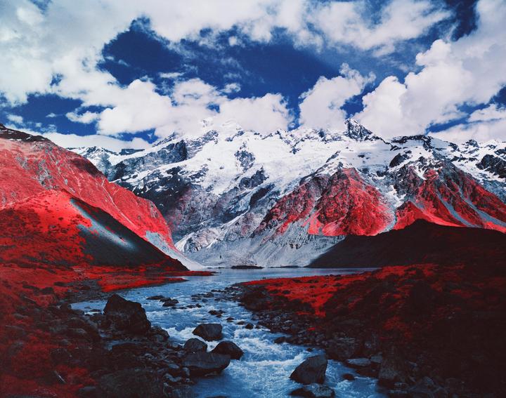 A river flows through a valley with red foliage and snow-capped mountains under a cloudy sky.