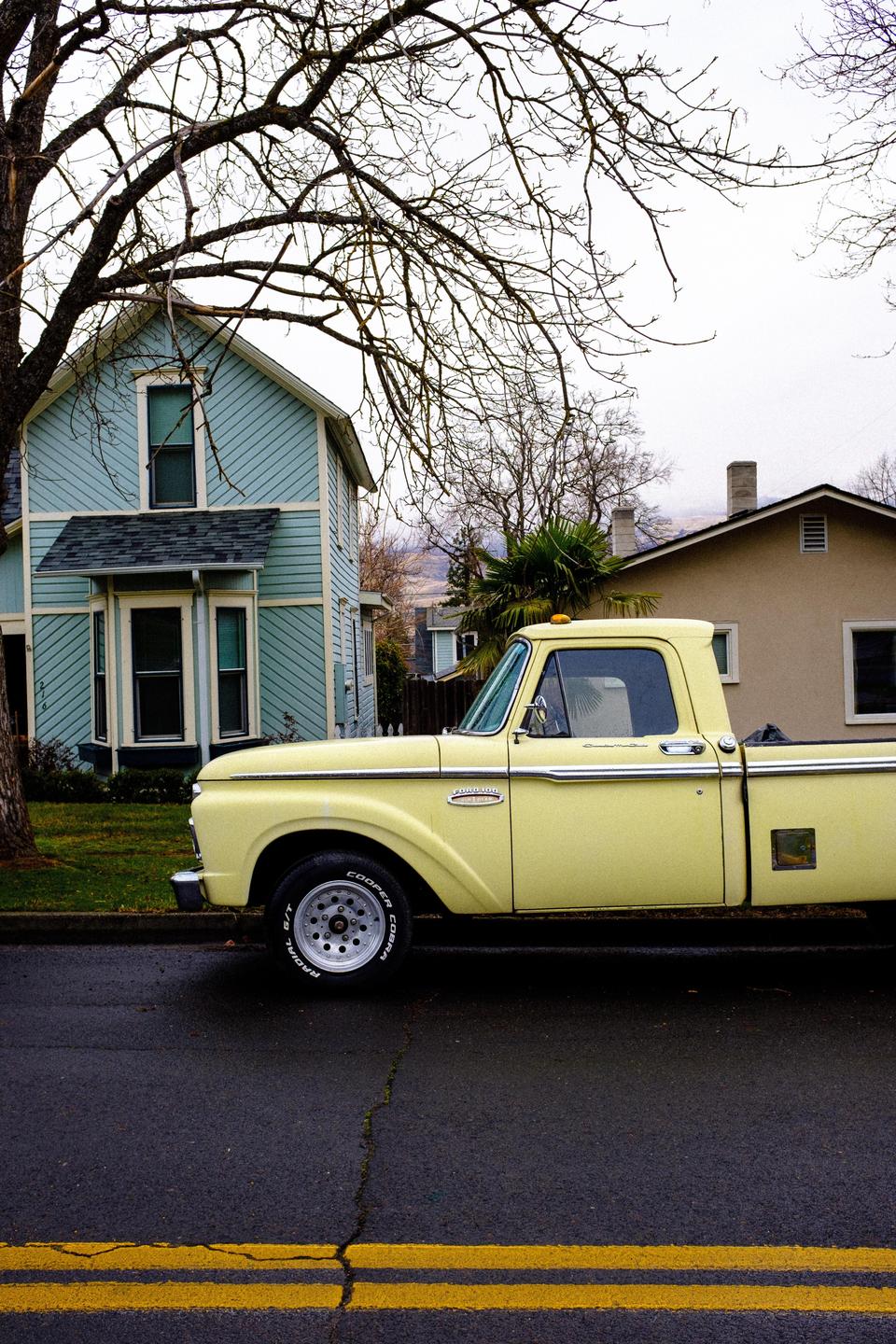 A yellow pickup truck is parked on a street in front of a light blue house with a bare tree in the foreground.