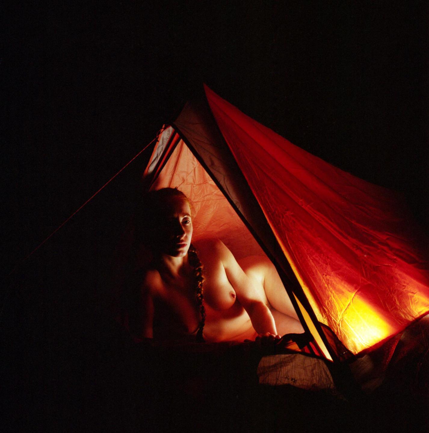 A woman with a braided pigtail lies inside a tent illuminated by a warm light at night.