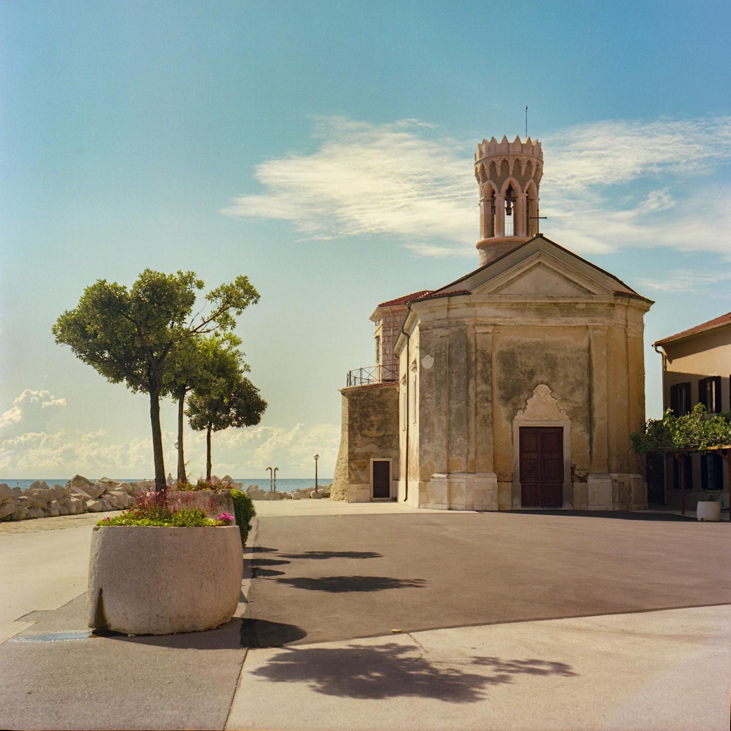 A church with a bell tower stands next to a paved area with trees and a planter, with the sea and a rocky shore in the background.