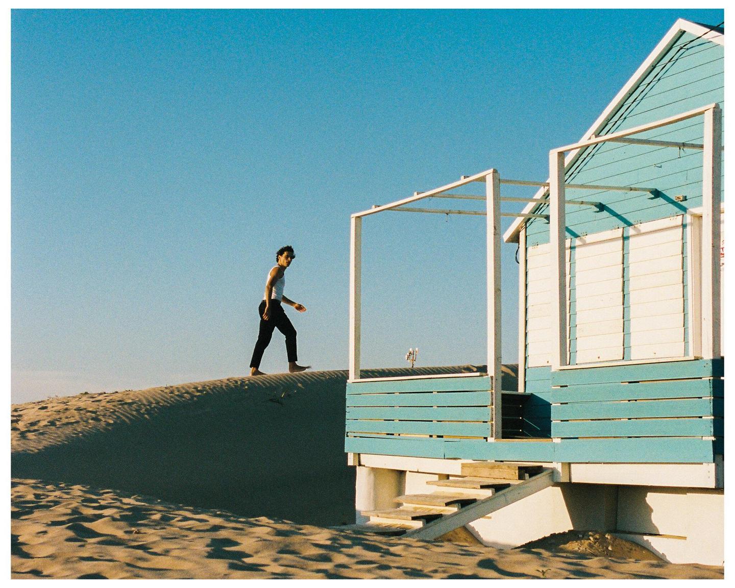 A man walks barefoot on a sand dune next to a blue and white beach house.