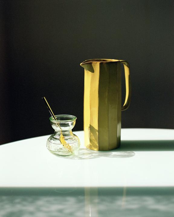 A yellow pitcher and a small glass vase with a stirrer are arranged on a white surface.