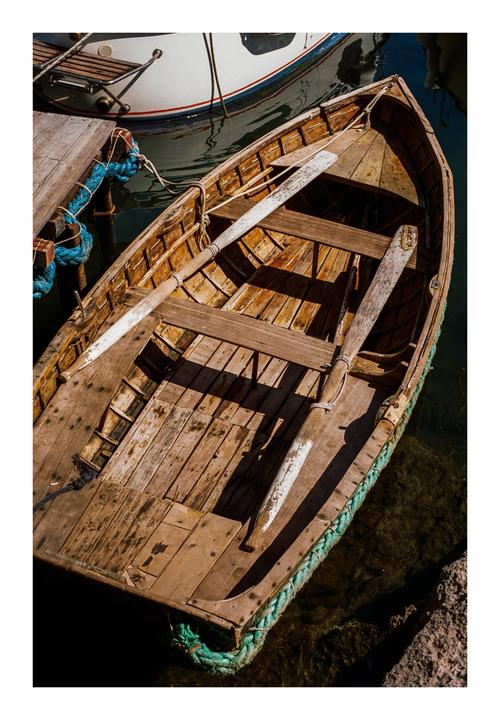 A wooden rowboat with oars rests in the water next to a dock and another boat.