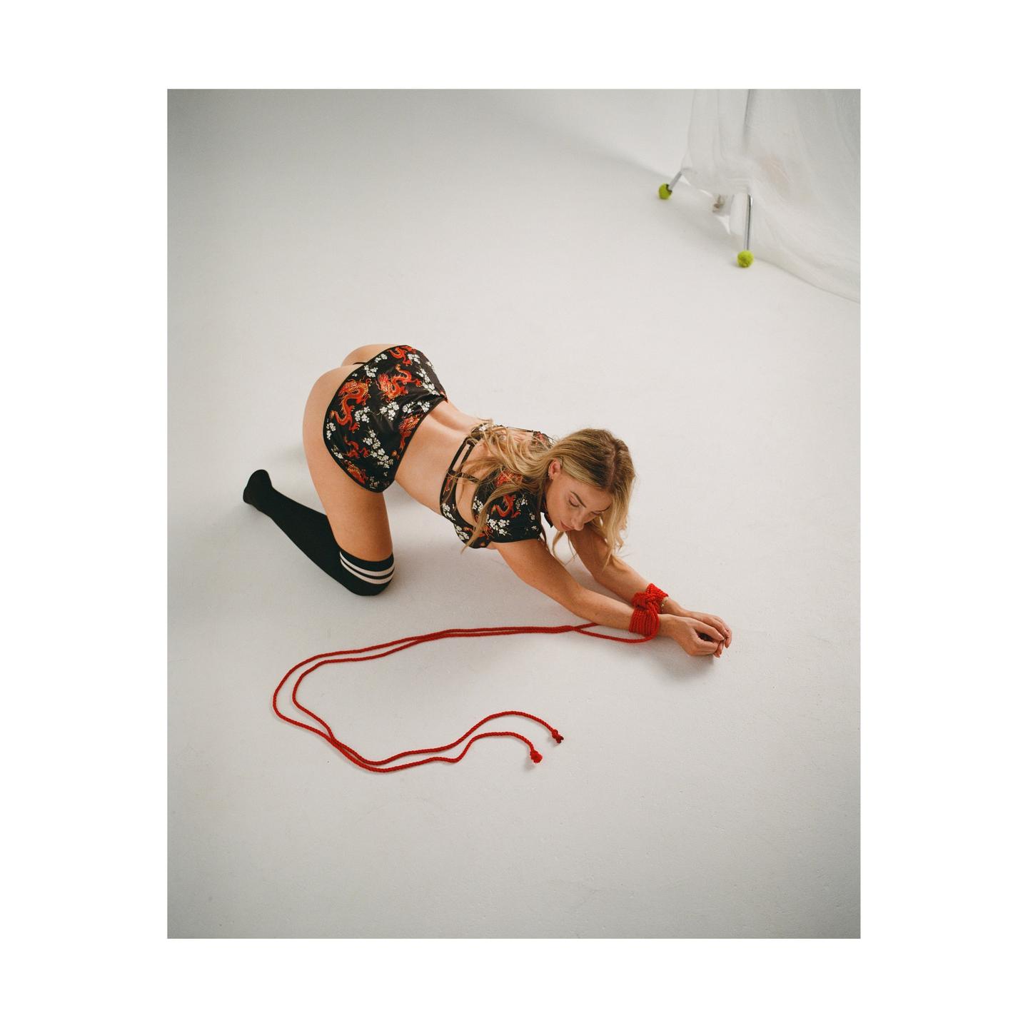 A woman in a floral top and shorts is on her hands and knees with her wrists bound by red rope.