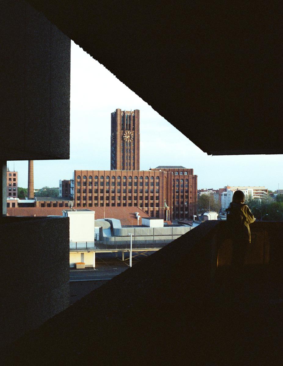 A woman stands on a dark balcony overlooking a city with a prominent clock tower.