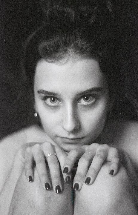 A woman with dark hair and dark nail polish looks directly at the viewer.