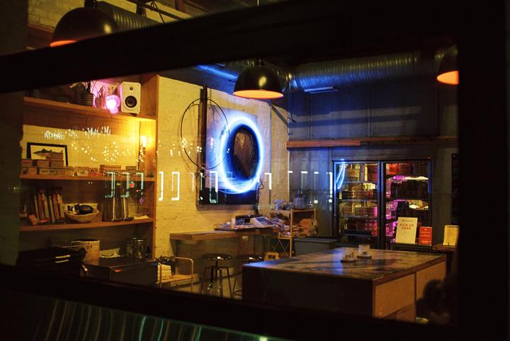 A cafe interior is visible through a window at night, featuring a glowing blue circular art piece and shelves with various items.