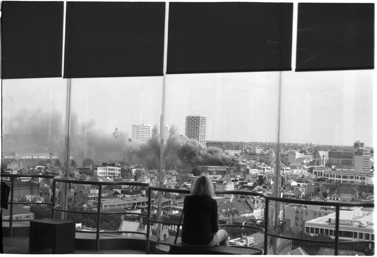 A woman sits on a balcony overlooking a city with smoke rising from a building.