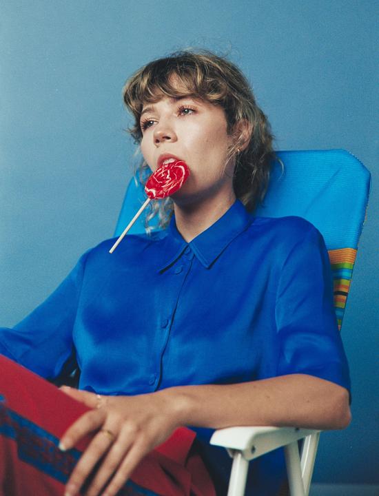 A woman with a lollipop in her mouth sits in a chair against a blue background.