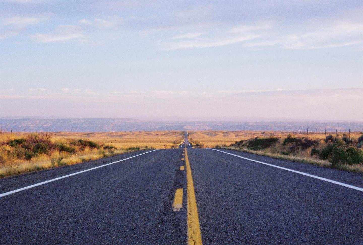 A long, straight road stretches into the distance through a dry, grassy landscape under a pale sky.
