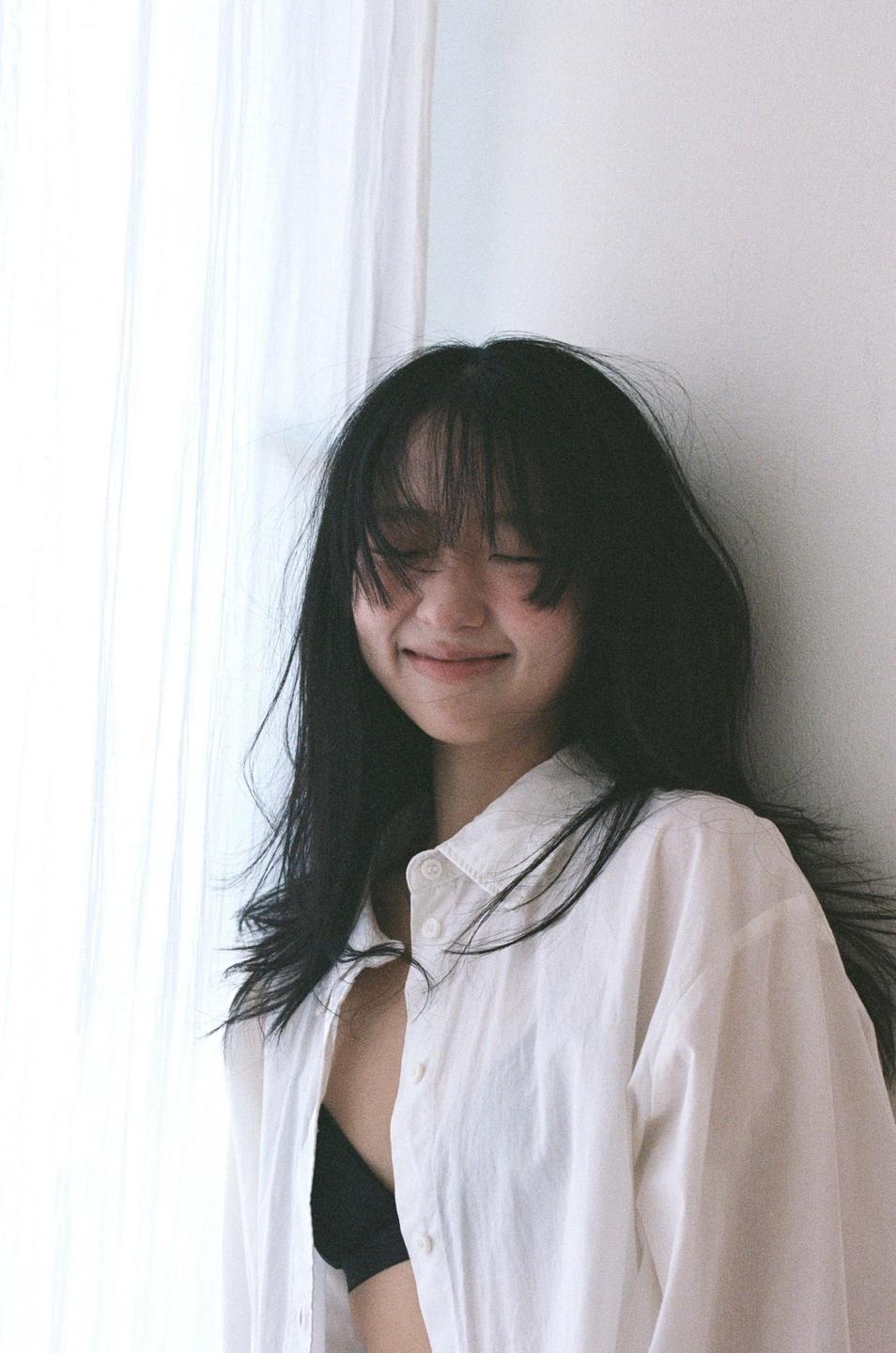 A woman with long dark hair and bangs smiles with her eyes closed, wearing a white shirt and black bra.