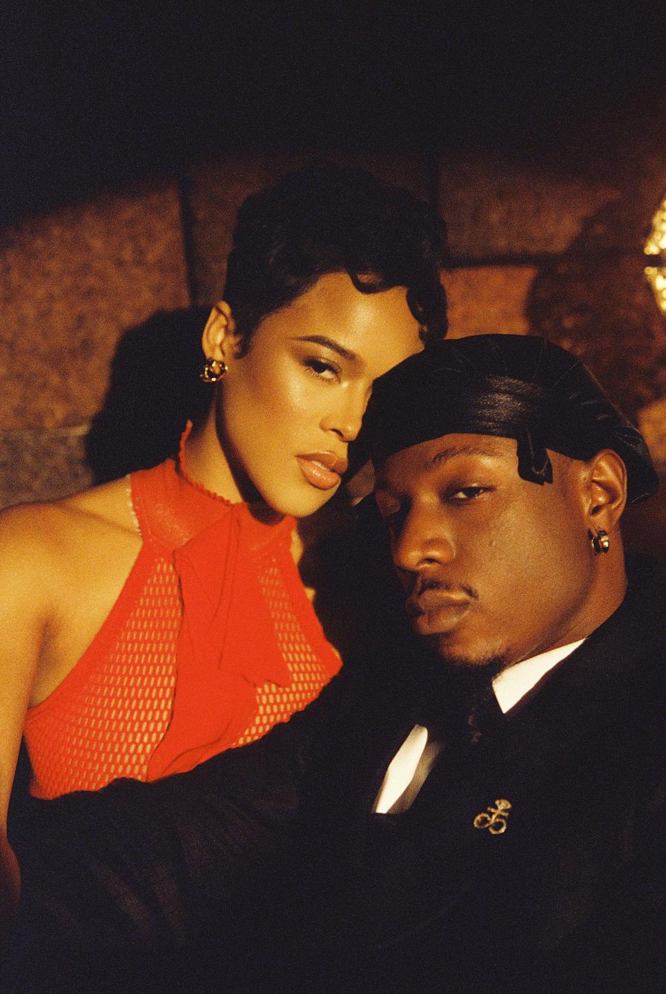 A woman in a red top and a man in a black suit and durag pose for a portrait.