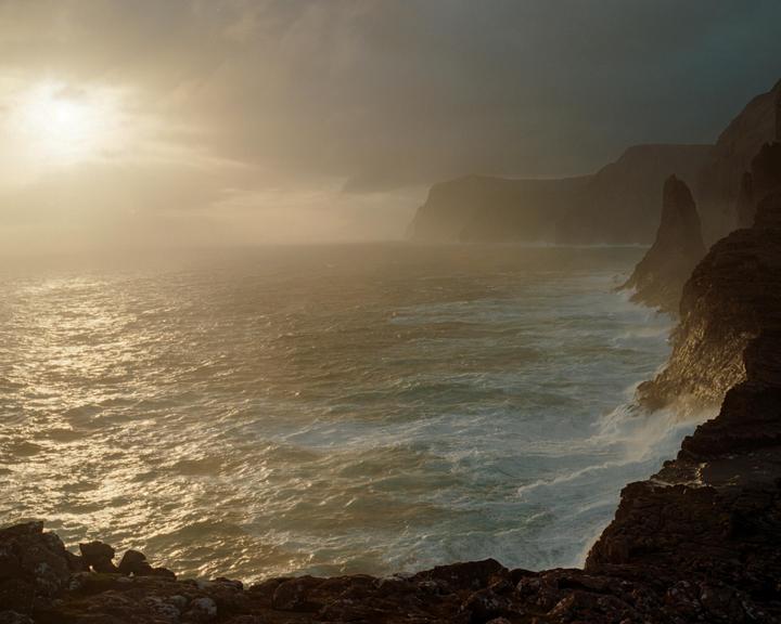 Waves crash against a rocky coastline under a hazy sky with a bright sun.