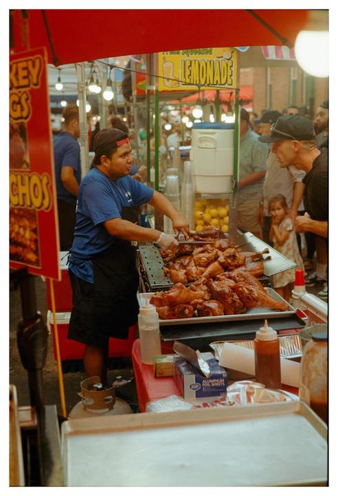A man in a blue shirt and black apron serves roasted turkey legs from a food stall at a market.