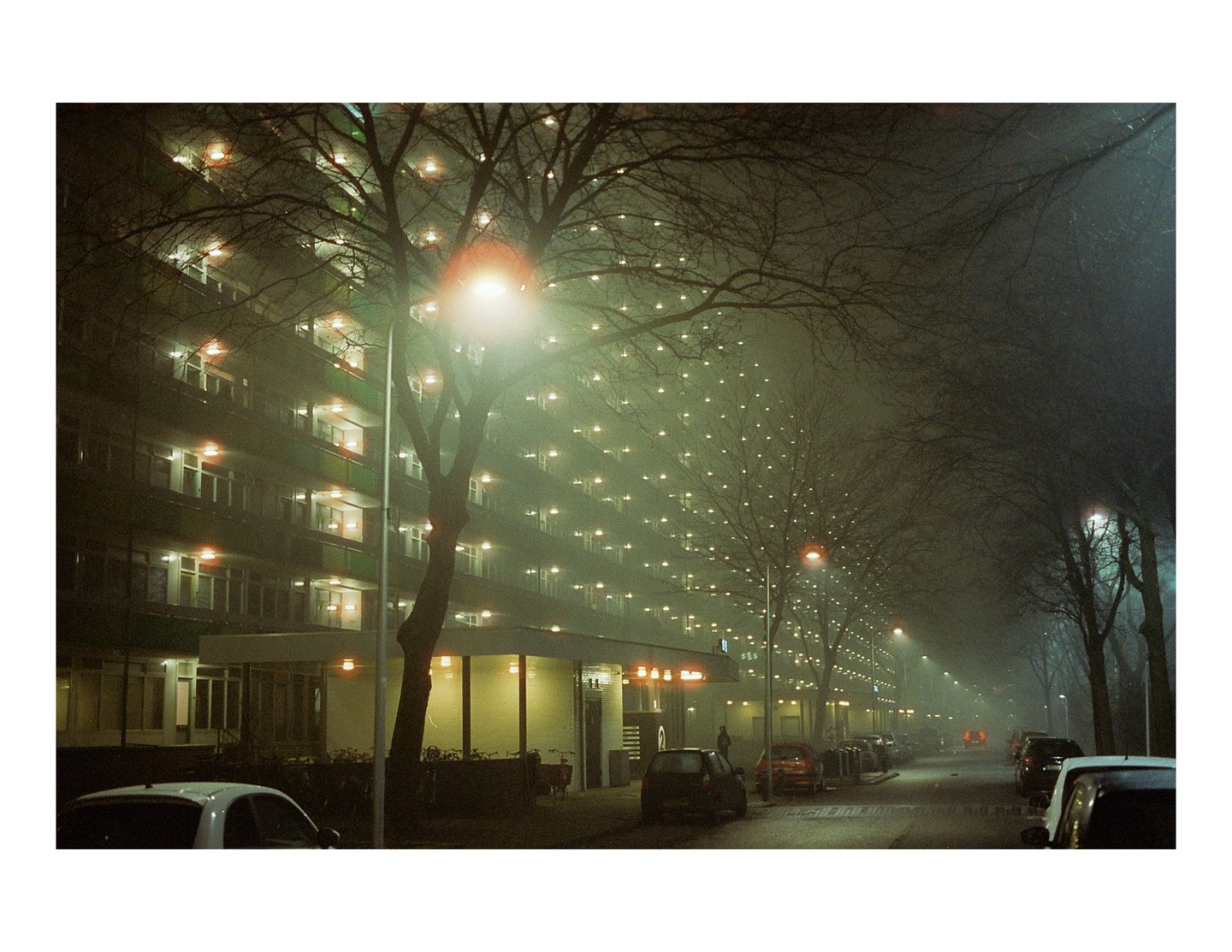 A street with parked cars and a large apartment building is illuminated by streetlights and interior lights on a foggy night.