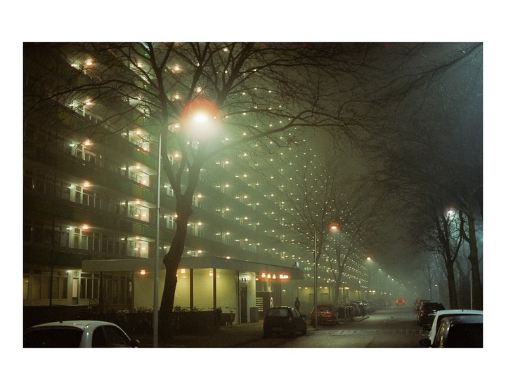 A street with parked cars and a large apartment building is illuminated by streetlights and interior lights on a foggy night.