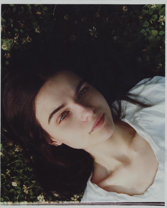 A woman with dark hair and a white shirt is lying on the ground surrounded by green foliage and small white flowers.
