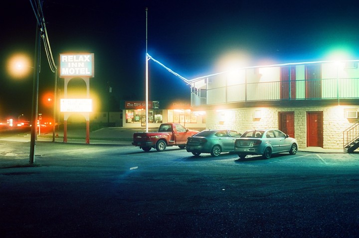 A motel sign and building are illuminated by neon lights at night, with several cars parked in front.