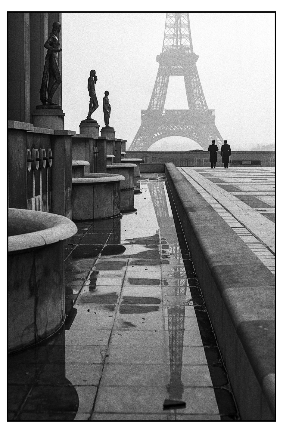 Two men walk towards the Eiffel Tower, which is reflected in a puddle on the ground, past a row of statues and fountains.