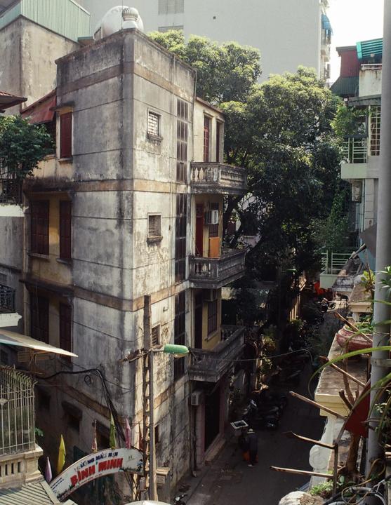 A narrow street is visible between two tall, weathered buildings with balconies and many trees.