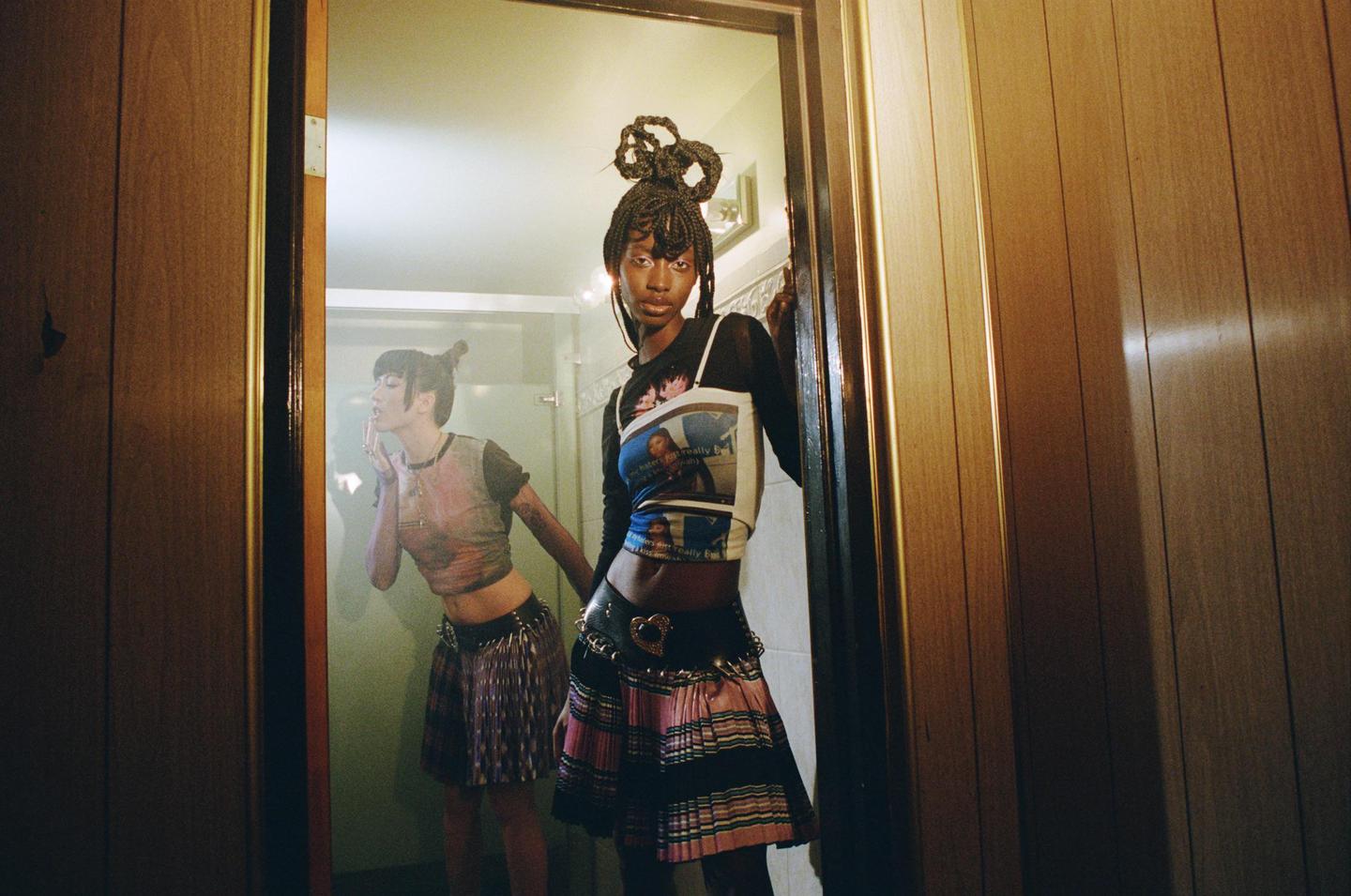 A woman with braided hair stands in a doorway, looking at her reflection in a mirror where another woman is visible.