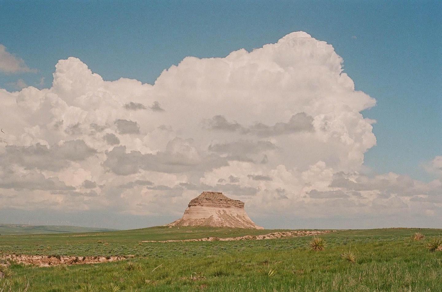 A large, flat-topped hill stands in a green field under a sky filled with cumulus clouds.
