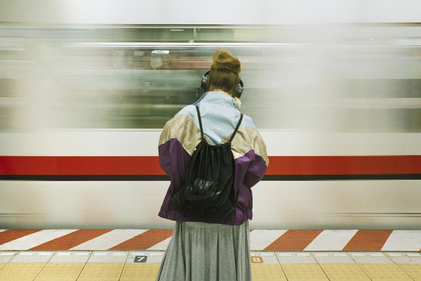 A woman with headphones and a backpack stands on a subway platform as a train passes by.